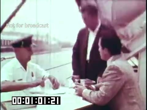 The still from 'Immigrant Heritage' features a scene on a boat or dock, where a group of men engages in conversation. A uniformed officer is seated at a table with documents, while two other men, one in a suit and another in a formal attire, stand nearby. The setting suggests a moment of immigration processing or a discussion related to heritage, aligning with the film's educational theme about immigrant experiences, particularly reflecting on Irish heritage. The visual tone is likely vibrant, characteristic of the film era.