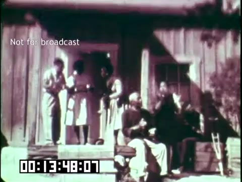 The still from 'Immigrant Heritage' features a group of people gathered on the porch of a wooden building. They appear engaged in conversation or activity, with some holding objects, suggesting a cultural gathering. The setting implies a historical context related to immigration and heritage, possibly highlighting community and cultural connections. The timecode indicates a specific moment in the film, while the watermark notes it is not for broadcast.