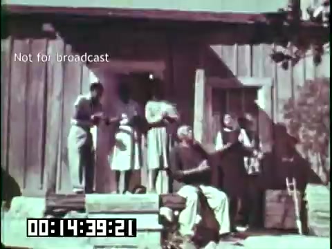 The still from 'Immigrant Heritage' features a group of individuals gathered outside a rustic wooden building. They appear to be engaged in musical activity, with some holding instruments. The scene evokes a sense of community and cultural expression, aligning with the film's theme of celebrating immigrant heritage. The overall atmosphere is warm and inviting, reflecting a connection to family and tradition.
