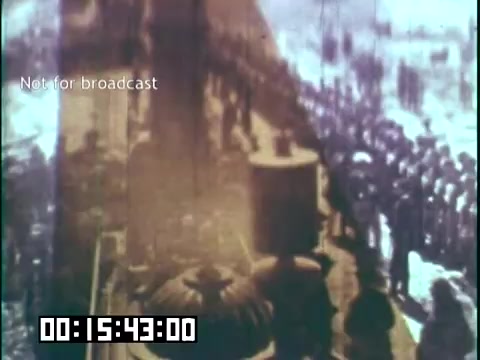 The still from 'Immigrant Heritage' features a historical scene depicting a train platform where a large group of people, likely immigrants, is gathered. The image captures a steam locomotive, with visible smoke and steam, emphasizing the transportation theme. The crowd appears engaged, perhaps awaiting the arrival or departure of the train, reflecting the era's migration experiences. The color tone suggests it may be an older film, adding to its historical context. The timestamp indicates the scene is part of a broader narrative on immigrant heritage.