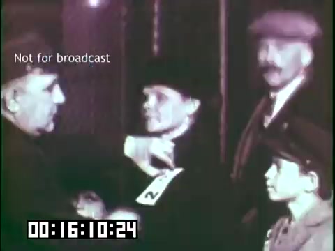 The still features a scene from the educational film 'Immigrant Heritage,' associated with the Irish Rovers. It depicts a moment of arrival, likely at an immigration checkpoint. A man in official attire interacts with a woman holding a numbered identification card, while another man and a young boy observe. The setting conveys the emotions and challenges faced by immigrants. The shot is framed in soft focus, emphasizing the characters' expressions and the gravity of the moment.