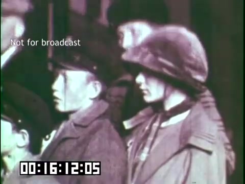 The still from 'Immigrant Heritage' features a group of individuals, likely immigrants, dressed in early 20th-century clothing. One person is wearing a military-style helmet, while another has a cap and appears younger. The scene seems to capture a moment of contemplation or waiting, perhaps at an immigration point or public event, highlighting themes of heritage and identity. The visual style is indicative of old film, contributing to the educational tone of the project. The watermark indicates it's not for broadcast.