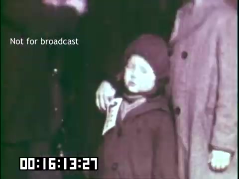 The still from 'Immigrant Heritage' features a young child dressed in a coat and hat, standing between two adults. The child appears to be holding a document or identification card. The image is in muted colors, reflective of its historical context. The scene captures a moment likely related to immigration, emphasizing the themes of heritage and identity. A timestamp and a watermark indicate the still is not for broadcast.