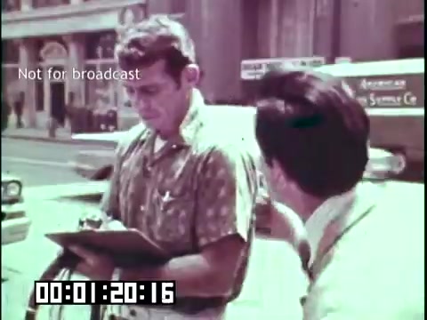 The still from 'Immigrant Heritage' features two men in a city setting. One man, wearing a patterned short-sleeve shirt, is holding a clipboard and appears to be engaged in a conversation with the other man, who is dressed in a lighter-colored shirt. The background includes urban scenery, indicative of a bustling environment, possibly suggesting a scene related to gathering information or interviews. The text 'Not for broadcast' indicates that this footage is intended for internal use only.