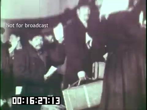 The still from 'Immigrant Heritage' features a group of men, some appearing weary, gathered in a setting that suggests they are immigrants. They carry luggage and wait in line, likely during a processing moment upon arrival. The black-and-white image conveys a sense of historical significance, capturing the immigrant experience. A timestamp is visible in the corner, indicating its position in the film.