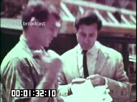 The still from 'Immigrant Heritage' features two men engaged in conversation, likely discussing topics related to immigration. One man appears to be examining a document or paper, while the other observes closely. The setting suggests an informal yet significant interaction, possibly highlighting cultural exchange or personal stories associated with immigrant experiences. The overall tone is educational, reflecting the film's focus on heritage. The image captures a moment of dialogue, emphasizing the importance of communication in understanding immigrant backgrounds.