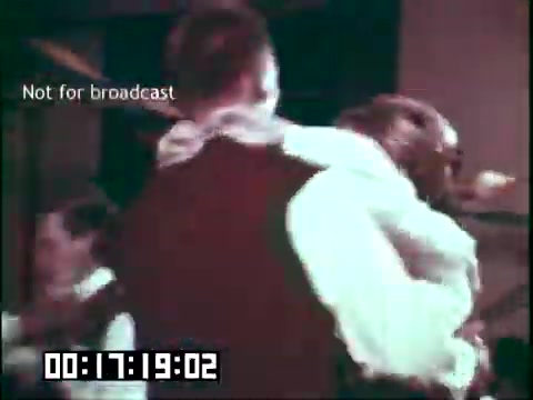 The still from the educational film 'Immigrant Heritage' featuring the Irish Rovers showcases a lively scene, likely depicting a dance or cultural celebration. Participants in traditional attire are engaged in an animated activity, emphasizing the vibrant customs associated with Irish heritage. The image includes a timestamp and a watermark indicating it's not for broadcast, suggesting it is either a behind-the-scenes look or a non-commercial preview of the film.