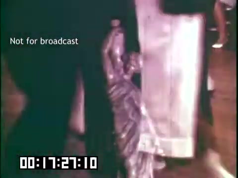 The still from 'Immigrant Heritage' features a close-up of a statue resembling the Statue of Liberty, possibly being held or carried. It captures an atmosphere of celebration or cultural significance, reflecting themes of immigration and heritage. The surrounding area shows blurred figures, hinting at a community gathering, while the timestamp and 'Not for broadcast' label indicate it is part of a larger educational film.