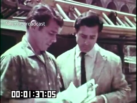 The still from 'Immigrant Heritage' features two men engaged in conversation, with one holding a piece of paper, possibly discussing important documents or information related to immigration. The setting appears to have a casual atmosphere, indicated by their clothing styles. The film likely aims to explore themes of heritage and the immigrant experience, possibly through personal stories or historical context. The timestamp indicates it's focused on a specific moment in the narrative.