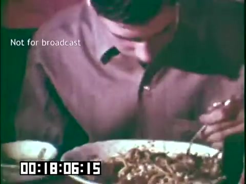 The still from 'Immigrant Heritage' features a man focused on his meal, which appears to be a sizeable portion of noodles. He is seated at a table, dressed in a button-up shirt with a collar, and is using a fork to eat. The image conveys a sense of cultural exploration through food, highlighting aspects of immigrant experiences. The timestamp and 'Not for broadcast' watermark indicate it is a protected showing from the film.