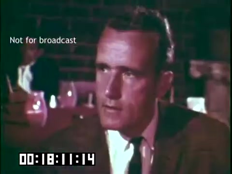 The still from the educational film 'Immigrant Heritage' features a man in a suit with a serious expression, engaged in conversation. The background includes dimly lit tables set with glasses. The image is marked 'Not for broadcast' and has a timestamp indicating it is from a segment of the film. The overall tone suggests a thoughtful or reflective moment, likely discussing themes related to Irish heritage.