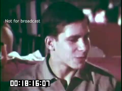 The still from the educational film 'Immigrant Heritage' featuring the Irish Rovers shows a young man engaged in conversation. The setting appears to be relaxed, possibly in a social or community space. The image is marked 'Not for broadcast' and includes a timestamp in a lower bar. The lighting is soft, suggesting a warm atmosphere, fitting the themes of cultural exploration and storytelling associated with the Irish Rovers.