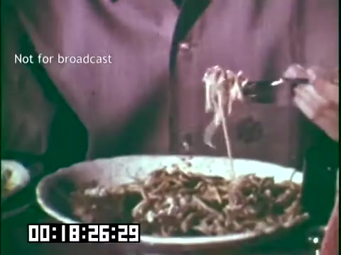 The still from 'Immigrant Heritage' features a close-up of a plate of food, likely showcasing a cultural dish associated with immigrant heritage. A person's hand is seen using a fork to lift noodles or strands of food from the plate. The background is blurred, emphasizing the food, and there is a time code at the bottom. The still also includes a watermark stating 'Not for broadcast.'