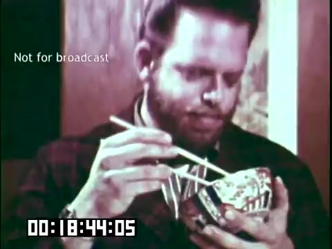 The still from 'Immigrant Heritage' features a man with a beard holding a decorative bowl while using chopsticks to eat. The scene highlights cultural practices, likely emphasizing the diversity within immigrant communities. The visual suggests a focus on shared experiences and the integration of different cultural traditions. The timecode indicates a specific moment in the film, and the label 'Not for broadcast' suggests it is meant for an educational context rather than public airing.