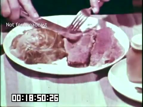 The still from 'Immigrant Heritage' features a close-up of a plate of food being served. A hand is using a fork and knife to cut into a piece of meat alongside a serving of cabbage. The table setting includes a small jar, possibly for condiments. The scene captures a moment emphasizing traditional Irish cuisine, likely reflecting the cultural aspects of immigration. The timecode indicates it is a specific moment in the film, which aims to educate about immigrant heritage.