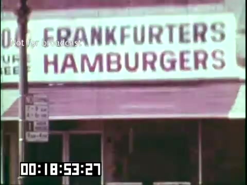 The still presents a storefront sign that reads 'FRANKFURTERS HAMBURGERS,' indicating a food establishment. The film context suggests a focus on immigrant heritage, possibly showcasing the cultural influences in the culinary landscape. The visual style aligns with vintage 16mm footage, emphasizing a historical perspective on immigrant experiences.
