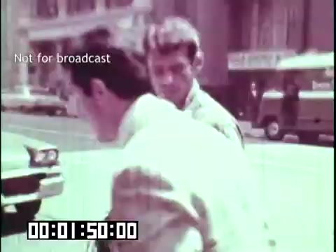The still from 'Immigrant Heritage' shows two men interacting on a city street. One man is facing away, dressed in a light-colored suit, while the other is in close proximity, observing him. The scene captures a moment of casual street activity, with vintage cars and buildings in the background, indicative of a mid-20th century urban setting. The time indicator suggests it is at the 1 minute and 50 second mark of the film. Overall, it reflects the social context of immigrant experiences.