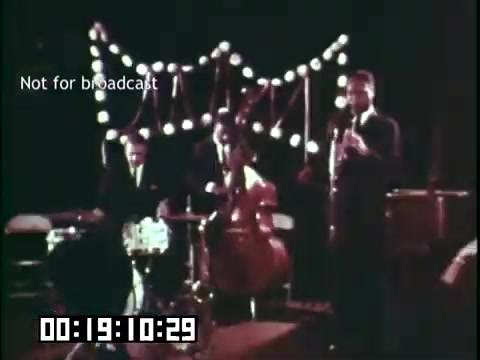 The still from 'Immigrant Heritage,' featuring the Irish Rovers, depicts a live musical performance. Three musicians are on stage: a drummer, a cellist, and a clarinet player, creating a lively atmosphere. The background is adorned with decorative lights, enhancing the festive feel of the scene. The image carries a timestamp and a notice indicating it is 'Not for broadcast.'