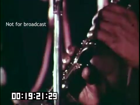 The still from 'Immigrant Heritage' shows a close-up of a person's hand playing a musical instrument, likely a trumpet or similar brass instrument, emphasizing the connection between music and cultural heritage. The frame includes a timestamp and a 'Not for broadcast' label, suggesting it is part of an educational film that highlights the role of music in immigrant experiences. The focus on the hands conveys passion and skill in the performance.