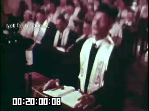 The still from 'Immigrant Heritage' features a congregation seated in a church setting, with a focus on a male figure in religious attire at the forefront. He is reading from a book, possibly a scripture, while wearing a ceremonial sash. The audience behind him appears engaged, suggesting a moment of worship or reflection. The visual context emphasizes themes of cultural heritage and community.