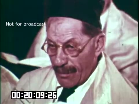 The still features an older man with a thoughtful expression, wearing glasses and a hat, seated among others. The setting appears to be a gathering related to the theme of immigrant heritage, possibly during a performance or discussion. The scene captures a moment of contemplation, reflecting the cultural themes explored in the film 'Immigrant Heritage.' The timestamp indicates it occurs around 20 minutes into the film, suggesting a key moment in the narrative.