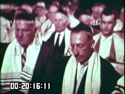 The still from 'Immigrant Heritage' depicts a group of men in formal attire, seated and engaged in a solemn gathering. They are wearing prayer shawls (tallit), indicating a religious or cultural ceremony. The atmosphere suggests a focus on tradition and community among immigrants, likely reflecting on their heritage. The setting appears to be indoors, possibly a synagogue or similar venue, emphasizing collective reflection or worship.