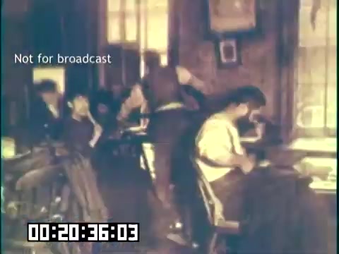 The still from 'Immigrant Heritage' features a group of individuals engaged in various activities in a historical setting. There's a focus on a man seated at a table, possibly writing or working, while others interact in the background. The scene captures a sense of community and the immigrant experience, highlighting cultural heritage. The image is muted in color, suggesting an older film quality, and includes a timestamp indicating its position in the film. The text 'Not for broadcast' is overlaid, indicating it is a private or protected still.