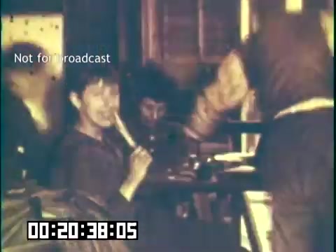 The still from 'Immigrant Heritage' features a group of individuals in a dimly lit setting. One person is playing a flute, while others are seated nearby, engaged in conversation or activities. The atmosphere suggests a communal or cultural gathering, reflecting the themes of heritage and connection explored in the educational film with the Irish Rovers. A timestamp indicates the specific moment within the film, and there's a watermark stating 'Not for broadcast.'