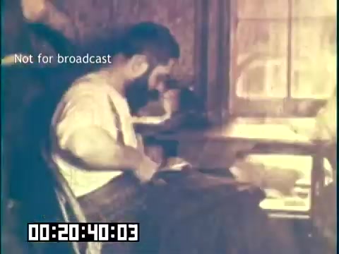 The still from 'Immigrant Heritage' features a man sitting at a table in a dimly lit room, engaged in an activity, possibly writing or reading. The setting evokes a historical context, likely portraying an immigrant's experience. The color tone suggests it may have been shot in a softer light, adding to the nostalgic feel of the film. Text indicating 'Not for broadcast' is superimposed, along with a timecode at the bottom.