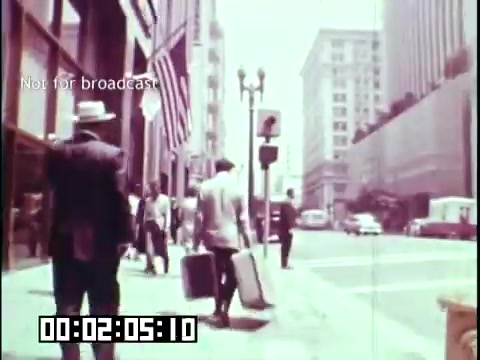 The still from 'Immigrant Heritage' features a bustling city street scene where individuals walk along the sidewalk, carrying suitcases. The presence of American flags suggests a theme of immigration and belonging. The vintage aesthetic and muted colors reflect the historical context of the film, which focuses on the experiences of immigrants, particularly highlighting their journey and adaptation in a new environment.