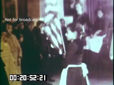 The 16mm still from 'Immigrant Heritage' featuring the Irish Rovers captures a moment likely during a cultural performance or commemorative event. In the foreground, a child holds flags, possibly symbolizing heritage or unity. The background has a diverse group of onlookers, indicating community engagement. The overall atmosphere conveys a sense of celebration and cultural pride. The image is labeled 'Not for broadcast,' suggesting it is intended for educational or archival purposes only.