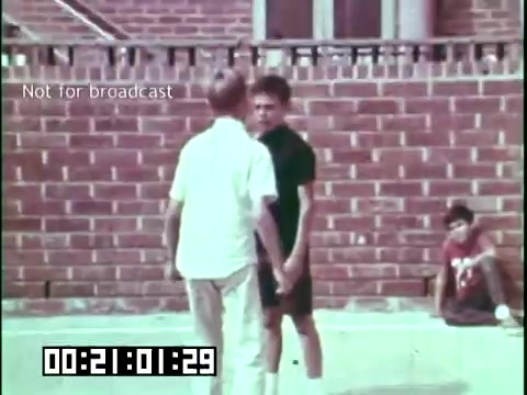 The still features two boys in a confrontation or playful interaction, set against a backdrop of a brick wall. One boy, wearing a white shirt, appears to be engaging with another boy in a black outfit. A third child is sitting in the background, seemingly observing the situation. The scene captures a moment of youth and perhaps the cultural dynamics depicted in the larger narrative of the educational film 'Immigrant Heritage' featuring the Irish Rovers. The film explores themes related to immigrant experiences and cultural identity.