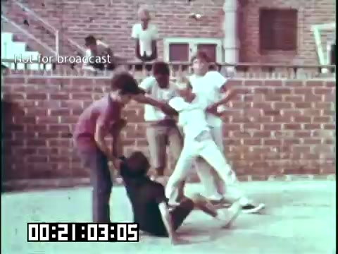 The 16mm still from 'Immigrant Heritage' features a group of children engaged in a playful interaction in an outdoor setting. The scene appears to depict a mix of fun and playful tussling among the kids, set against a brick wall backdrop. The environment suggests a community gathering place, indicative of the film's theme of cultural and social connections among immigrant communities. The overall tone conveys a sense of youthful energy and camaraderie.