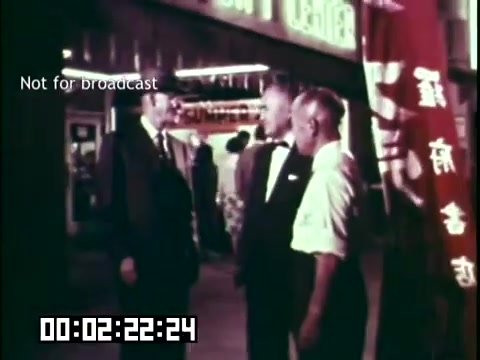 The still from 'Immigrant Heritage' features three men engaged in conversation, likely representing various cultural backgrounds. They stand in front of a building adorned with signs, indicating a vibrant community atmosphere. One man wears a suit, while another is in a white shirt, suggesting a formal gathering. The scene captures themes of diversity and interaction among immigrant communities, typical of the film's educational focus on heritage.