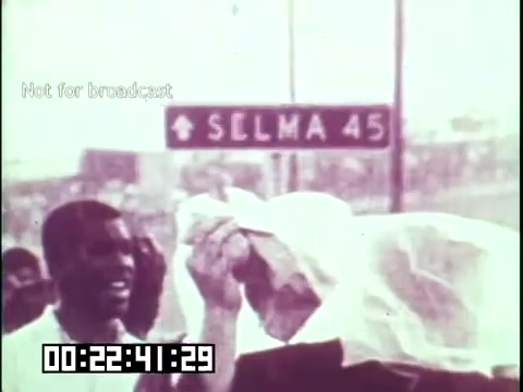 The 16mm still from 'Immigrant Heritage' features a scene likely related to a historical event, capturing individuals in a moment of reflection or struggle. One person appears to be wiping their forehead, possibly indicating the intensity of the situation or environment. In the background, a directional sign for Selma suggests a connection to the civil rights movement, emphasizing themes of social justice and heritage. The overall tone conveys a sense of perseverance and the impact of historical movements on immigrant experiences.