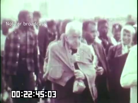 The still from 'Immigrant Heritage,' featuring the Irish Rovers, depicts a diverse group of people, including an elderly man wrapped in a blanket. The setting suggests a moment of arrival or gathering, likely reflecting themes of immigration and cultural heritage. The image has a vintage quality, typical of 16mm film, and conveys a sense of community and shared experience among immigrants.