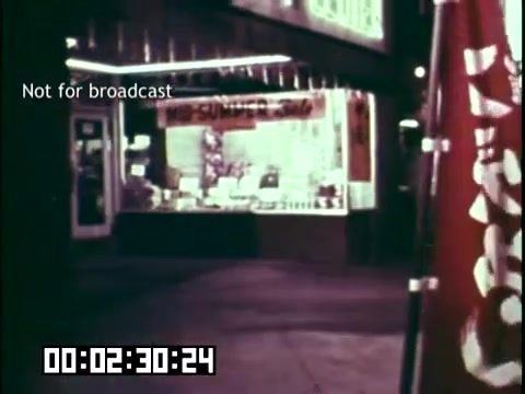 The still from 'Immigrant Heritage,' an educational film featuring the Irish Rovers, depicts a streetscape at night. A storefront is visible, illuminated with lights showcasing merchandise. The image exudes a nostalgic vibe, capturing an urban environment that reflects immigrant culture. In the foreground, a colorful flag stands, hinting at cultural pride or community representation. A digital timer in the corner indicates the film's progression. The note 'Not for broadcast' suggests it is for internal use only.