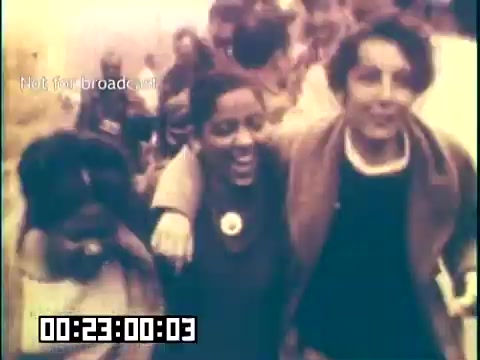 The 16mm still from 'Immigrant Heritage' features a group of people, with two individuals smiling and embracing each other. The background is filled with a crowd, suggesting a lively atmosphere. The color is likely muted or faded, characteristic of older film stock. The context hints at themes of cultural celebration and community, possibly exploring the Irish immigrant experience.