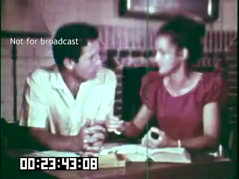 The still from 'Immigrant Heritage' features a man and a woman engaged in conversation. The setting appears to be a cozy indoor environment, likely a living room, with a visible fireplace. The man wears a light-colored shirt and the woman is in a red dress. They seem to be discussing or studying something, with books and notes in front of them. The frame includes a timestamp and a note indicating it's not for broadcast. The overall mood suggests a personal and educational exchange related to immigrant experiences.