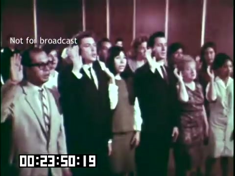 The still from 'Immigrant Heritage' features a diverse group of individuals standing together, raising their right hands, likely as part of a naturalization ceremony. The scene captures a moment of unity and solemnity, symbolizing their commitment to becoming citizens. The setting appears to be indoors, with a formal ambiance. A timestamp and 'Not for broadcast' label indicate the film format and context.