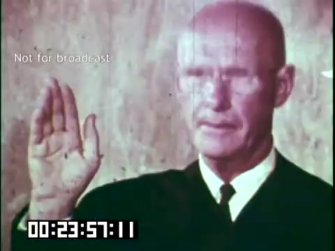 The still depicts a man in formal dress, likely a judge or official, raising his right hand in a gesture of oath. The background appears plain, emphasizing the figure. The image is likely part of a scene related to themes of immigration, possibly symbolizing commitment to a new country or heritage, aligning with the context of the educational film featuring the Irish Rovers. The timestamp suggests it is a moment captured during the film's runtime.