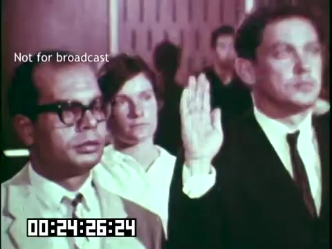 The still from 'Immigrant Heritage - Educational Film with the Irish Rovers' depicts a moment likely related to a naturalization ceremony. It features two men, one with glasses and a suit, and the other in formal attire, raising their right hands in an oath-taking gesture. A woman in the background appears to be observing the event. The setting is formal, possibly in a government building, reflecting themes of immigration and cultural heritage. The image is marked 'Not for broadcast,' indicating it is for educational use.