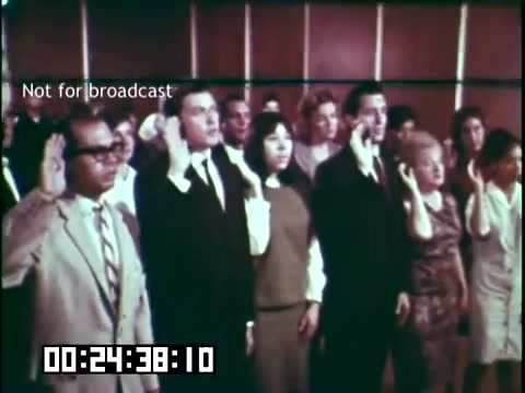 The still from 'Immigrant Heritage' features a diverse group of individuals standing in a hall, all with their right hands raised in a gesture of taking an oath. They appear to be participating in a naturalization ceremony or similar civic event. The setting has a formal atmosphere, indicated by the presence of a backdrop and a group of people dressed in business attire and casual clothing. In the lower corner, there is a timecode and a note indicating that the image is not for broadcast.