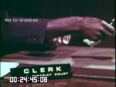 The still from the educational film 'Immigrant Heritage' features a close-up of a clerk's hand, performing an action at a desk with a nameplate reading “CLERK DISTRICT COURT.” The image conveys a sense of official business, likely related to legal or administrative proceedings, within the context of immigration themes explored by the film. The timestamp indicates it occurs at 24 minutes and 45 seconds into the film. The note 'Not for broadcast' suggests it is intended for educational use only.