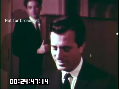 The still from 'Immigrant Heritage' features two men in a scene from an educational film. One man, dressed in a suit, is in the foreground with a focus on his expression. The second man is in the background, partially out of focus, suggesting a dialogue or interaction. The setting appears to be a formal or studio environment, indicated by the presence of soft lighting and a neutral backdrop. The timestamp at the bottom suggests it's from a specific point in the film.