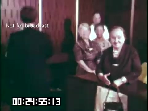 The still from 'Immigrant Heritage' features a scene likely in a public setting, such as a hall or community space. In the foreground, an older woman smiles while holding a handbag. Behind her, several individuals appear engaged in conversation or observation, contributing to a sense of community gathering. The atmosphere suggests a celebration or recognition of cultural heritage, relevant to the themes explored in the film. The timestamp indicates this moment occurs approximately halfway through the film.