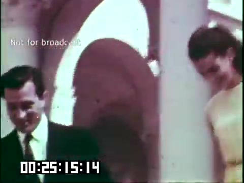 The 16mm still from 'Immigrant Heritage' featuring the Irish Rovers shows two individuals, a man and a woman, walking together. They appear to be in a formal setting, possibly an event or a gathering, with architectural columns in the background. The image captures a moment of camaraderie and highlights the cultural theme of the film. The timestamp suggests it occurs at a specific point within the documentary.