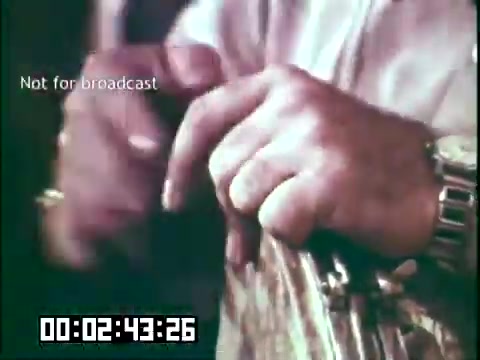 The still features a close-up of hands playing a musical instrument, likely indicating a moment of performance in the educational film 'Immigrant Heritage' featuring the Irish Rovers. The hands are focused on a fretboard, suggesting a connection to traditional Irish music. The timestamp and 'Not for broadcast' label indicate it's part of a film reel not intended for public airing. The overall context emphasizes the celebration of immigrant heritage through music.