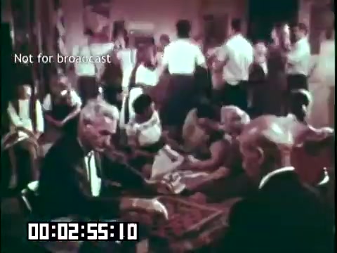 The still from 'Immigrant Heritage' features a gathering where a group of individuals is engaged in conversation and activities. In the foreground, two older men, dressed in formal attire, appear to be playing a game or engaging in a discussion. In the background, men and women are socializing, with some seated on the floor and others standing. The setting conveys a warm, communal atmosphere, likely reflecting elements of Irish culture as represented by the Irish Rovers in the film. The visual is rich in color and captures the essence of cultural celebration.