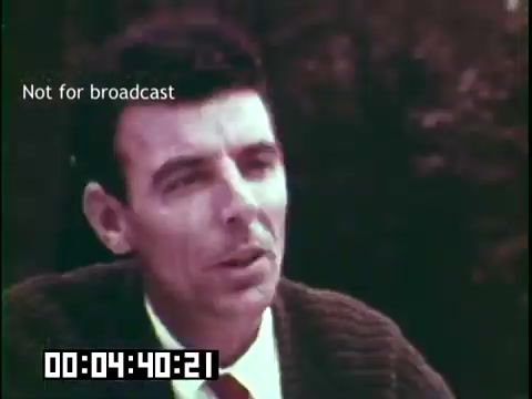 The still shows a man speaking, likely sharing insights related to immigration and cultural heritage. He is dressed in a sweater over a collared shirt and tie, suggesting a thoughtful or serious discourse. The background appears natural, possibly indicating a reflective or informal setting. The timestamp and 'Not for broadcast' label imply it's a segment from the film, focusing on the themes of immigrant experiences and heritage as explored by the Irish Rovers.